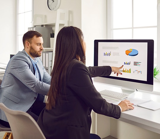 Two businesspeople analyzing data charts, graphs on pc monitor. Man and woman colleagues looking at graphs on computer screen preparing statistical report, discussing financial data in office